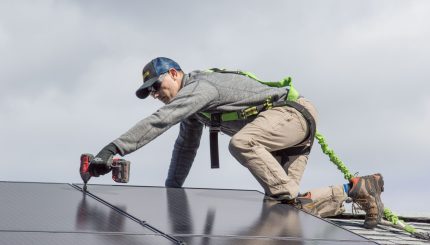 Man on roof of a building using a drill to install solar panels.